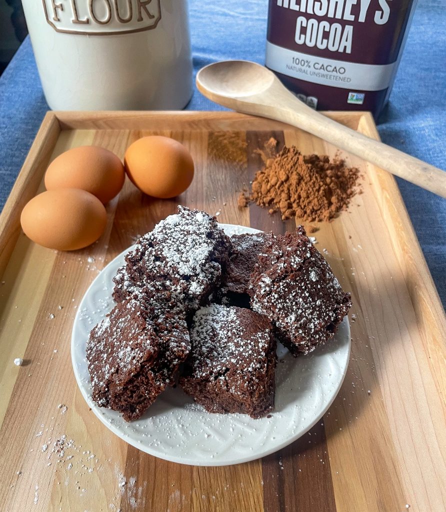 eggs, cocoa, and a finished batch of brownies on a plate sit atop a wooden tray with a wooden spoon resting on the side