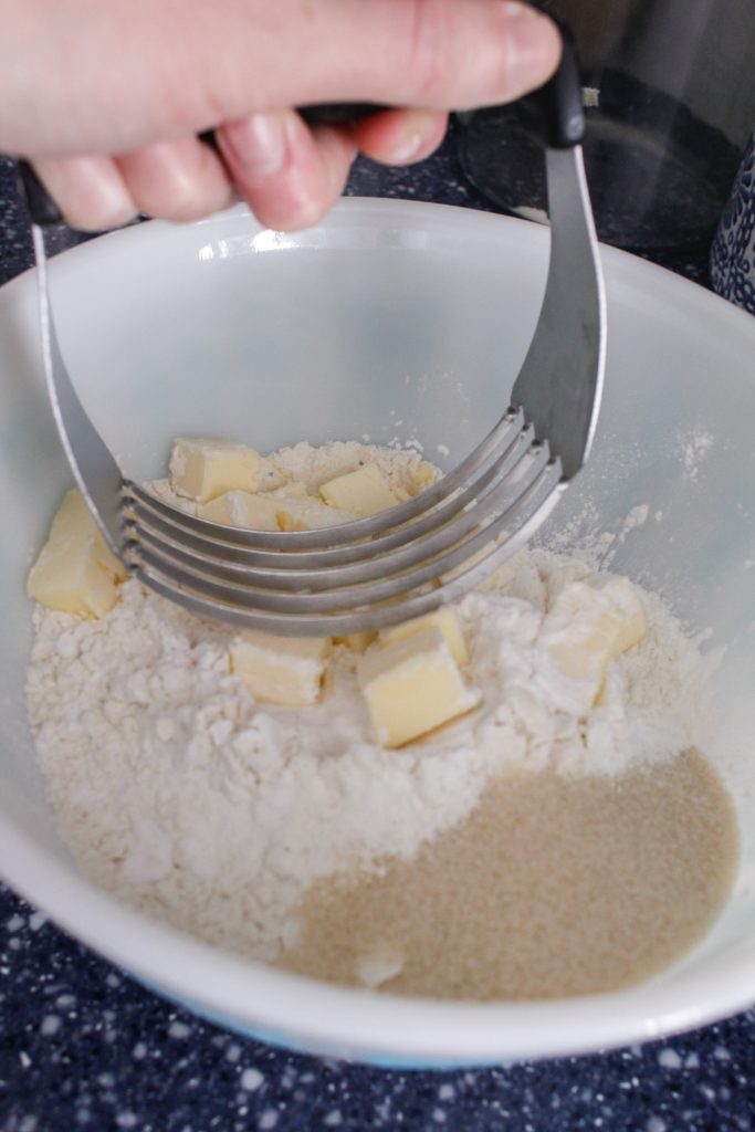 A pastry cutter mixes cubed butter, flour, and sugar in a bowl.