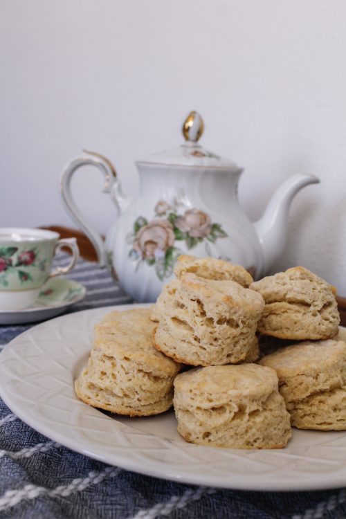 Scones sit on a white plate in front of a teapot.