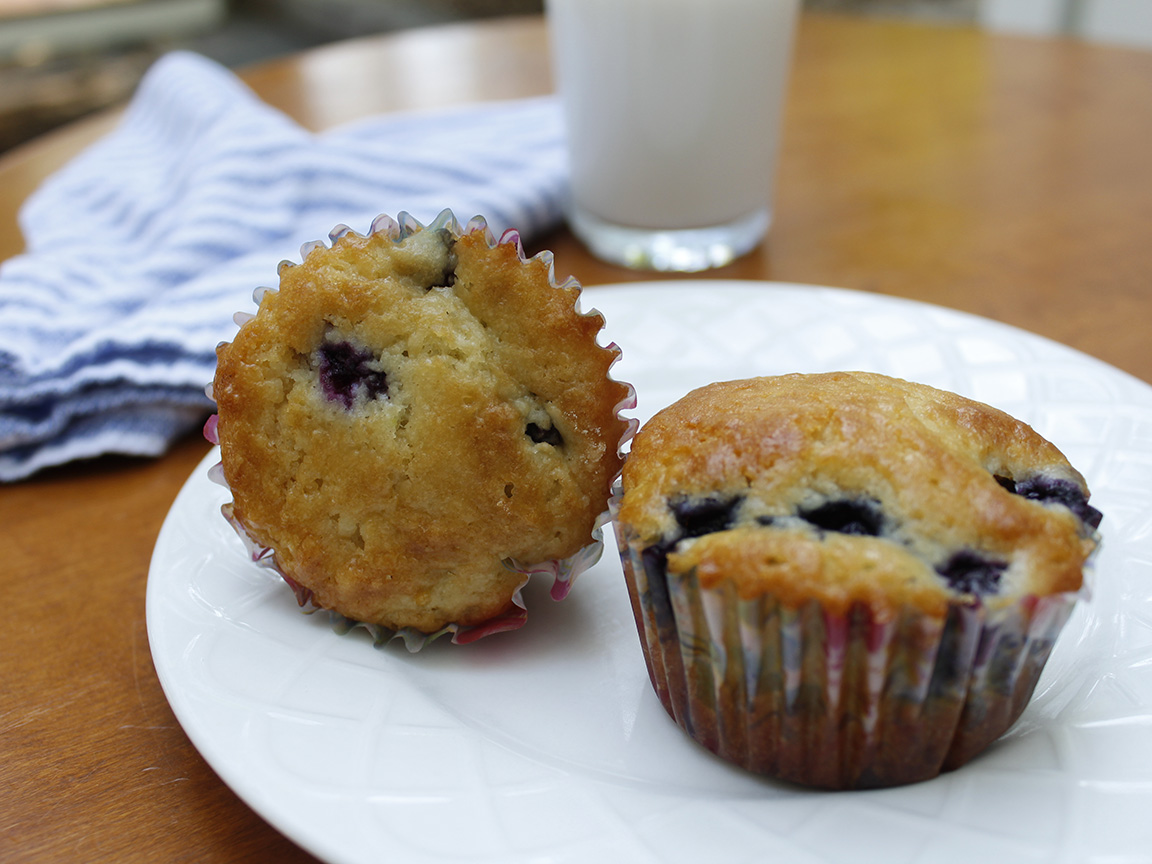 Two blueberry muffins sit on a white plate with a glass of milk in the background.