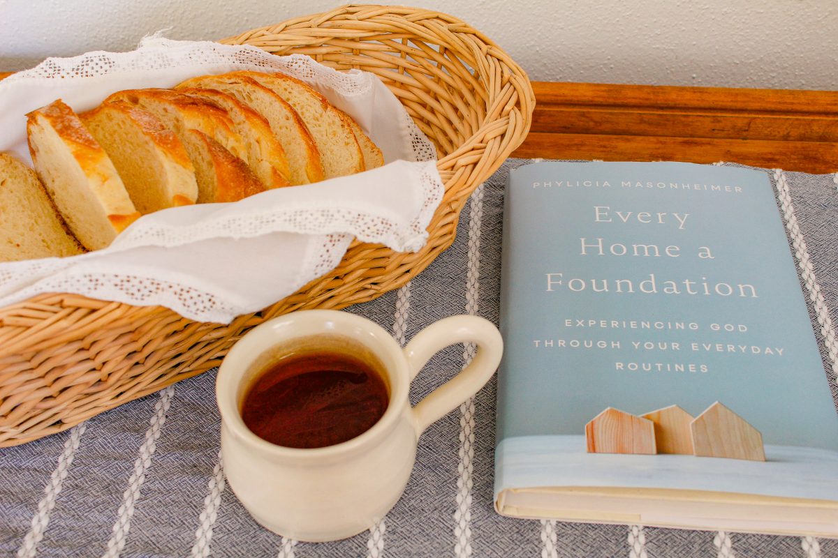 a cup of tea sits near a basket of bread and the book "Every Home a Foundation" by Phylicia Masonheimer
