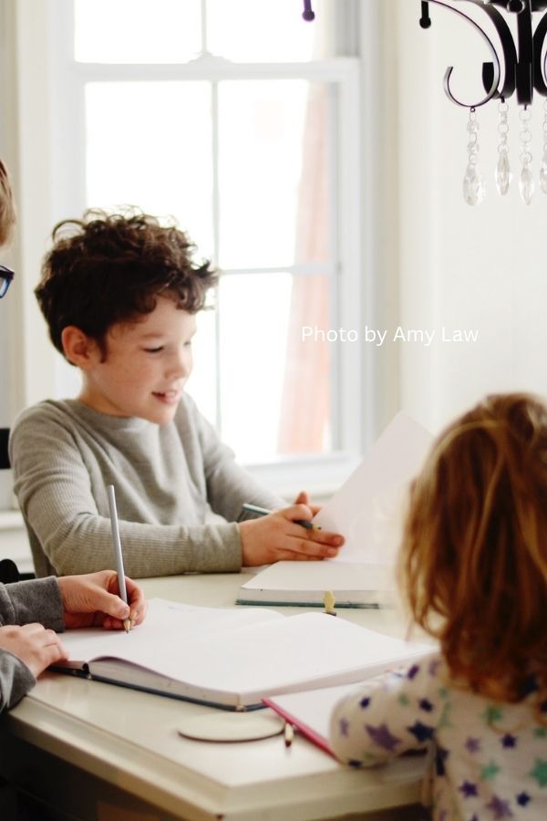 A boy and his siblings sit at a dining room table, happily doing their homeschool work.